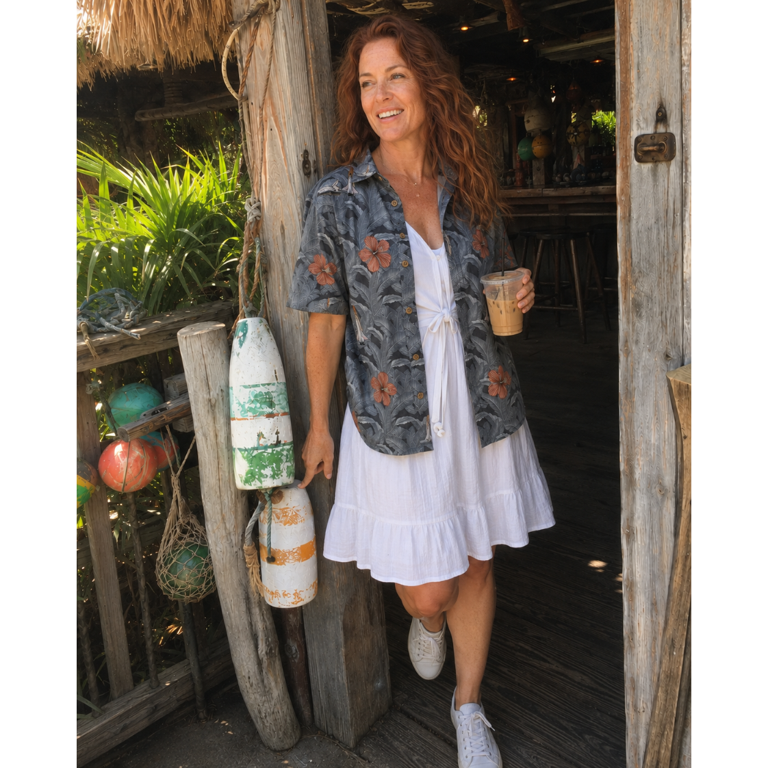 A woman stands in a rustic doorway wearing a white tiered ruffle dress with a gray floral short-sleeve button-up shirt layered over it, holding an iced coffee. She is posed next to a wooden post with hanging buoys.